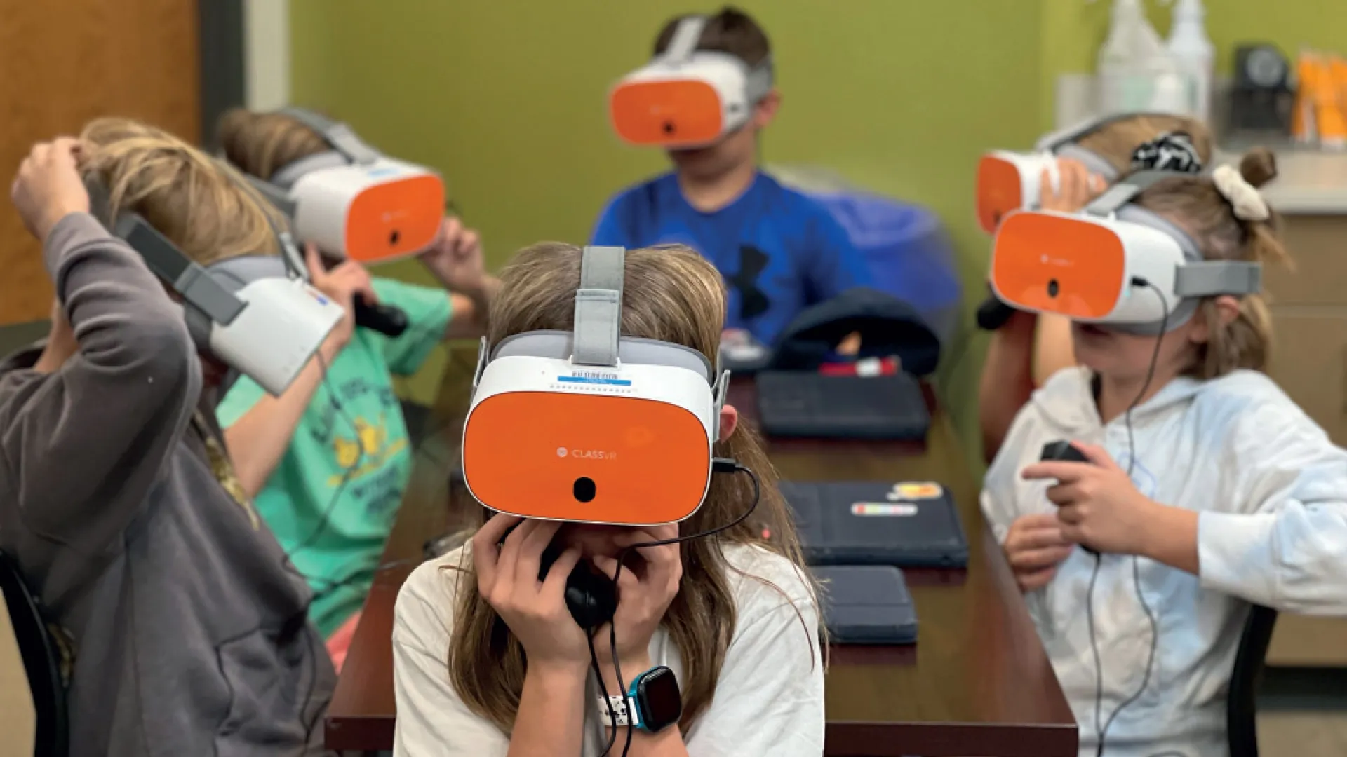 a picture of elementary school kids in a classroom, sitting around a table wearing oculus VR headsets
